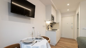 Una habitación compacta con una mesa de comedor preparada para dos personas, una kitchenette, un televisor de pantalla plana montado en la pared y una cama con cubrecama verde.