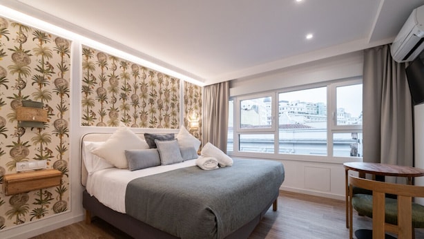 Un dormitorio moderno con una cama grande, una mesita de noche, una pequeña mesa y un ventanal con vistas a la ciudad.