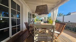 Une terrasse extérieure avec une table et des chaises en bois, un luminaire suspendu en osier et de grandes fenêtres.