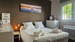 Une chambre à coucher avec un lit, des oreillers, une table de chevet avec une lampe, une commode, un tableau encadré au mur et une fenêtre donnant sur un paysage verdoyant.