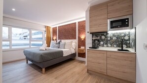 A modern bedroom with a bed, a wooden floor, a kitchen area with a microwave, and a window with curtains. - 306 — SB20 Boutique Studios | 1BR | 1BA | W/K (Madrid)