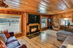 Ein Ferienhaus-Innenraum mit Kamin, Holzwänden, einer Couch und Blick auf eine verschneite Landschaft.