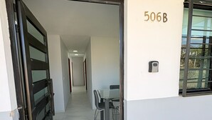 Un couloir mène à une salle à manger où se trouvent une table et des chaises, une porte noire et une fenêtre sur laquelle est inscrit le numéro 506B.