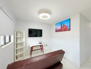 A room with a white wall, a mounted TV, a painting of a cathedral, a white desk with a computer, and a brown sofa. - Residencia Gran Coyula Orange 506B (Tonala)