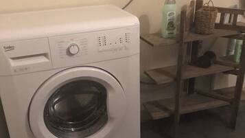 A front-loading washing machine with a control panel, a wicker basket, and a shelf with books.