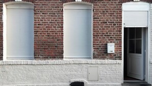A brick building with three closed roller shutters and a door. - Whole house in town, pets welcome (Caudry)
