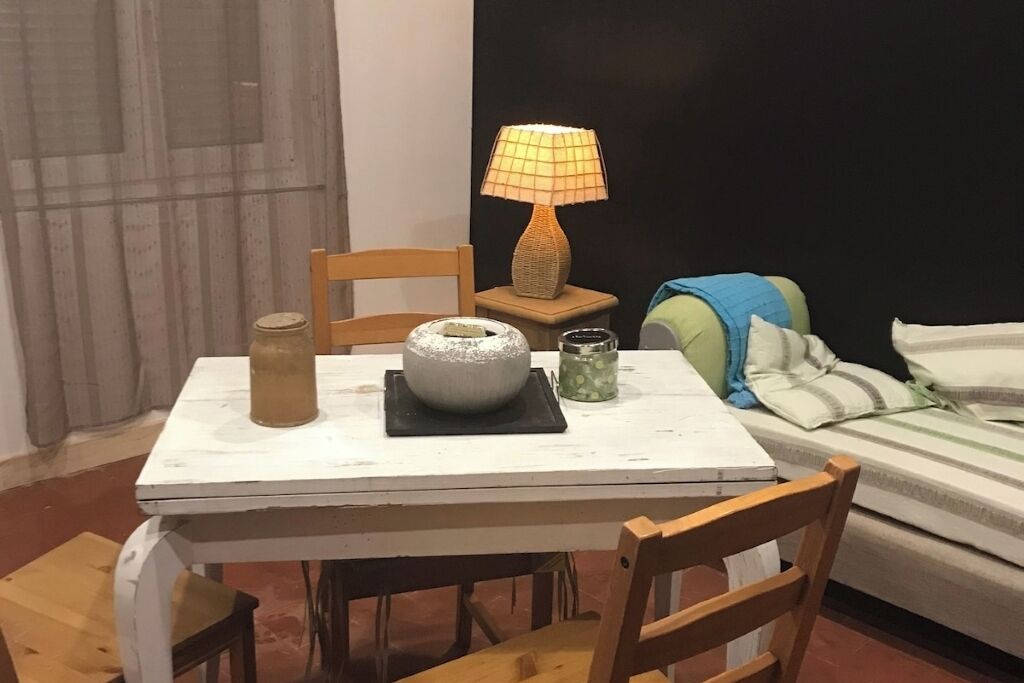 Un petit coin repas avec une table, des chaises, une lampe, un bocal et un lit avec des oreillers.