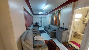 A living room with a sofa, a small table, and a washing machine. There is a door leading to a bathroom.