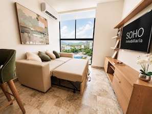 A modern living room with a sofa, a convertible bed, a wooden cabinet, and a large window offering a city view. - VITA 219 Apartment - Near the Sea by SOHO - 377A (Santa Marta)