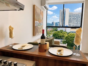 A dining table set with plates, napkins, and a wine bottle, with a cityscape view through a large window. - VITA 219 Apartment - Near the Sea by SOHO - 377A (Santa Marta)