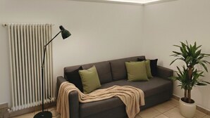 Ein Wohnzimmer mit einem dunkelgrauen Sofa, zwei grünen Kissen, einer beige farbenen Decke, einer Stehlampe, einer Heizung und einem Blumentopf.