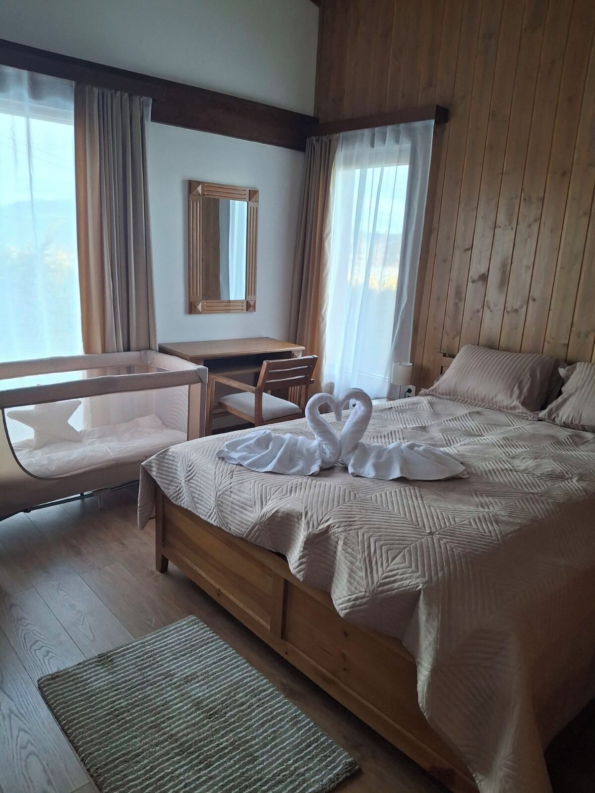 A hotel room with a wooden bed, a desk, a chair, and a large window with curtains.