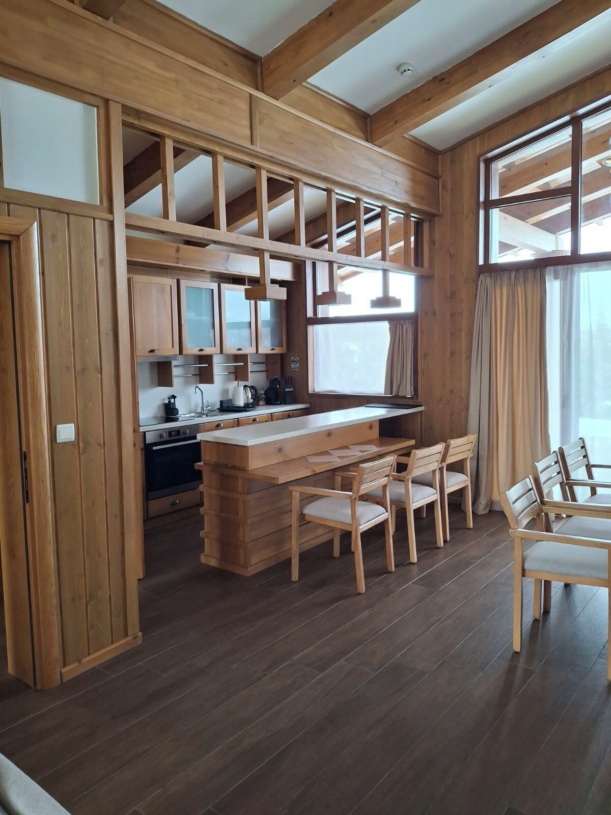 A spacious kitchen with wooden cabinets and a central island, a dining area with wooden chairs, and large windows providing natural light.