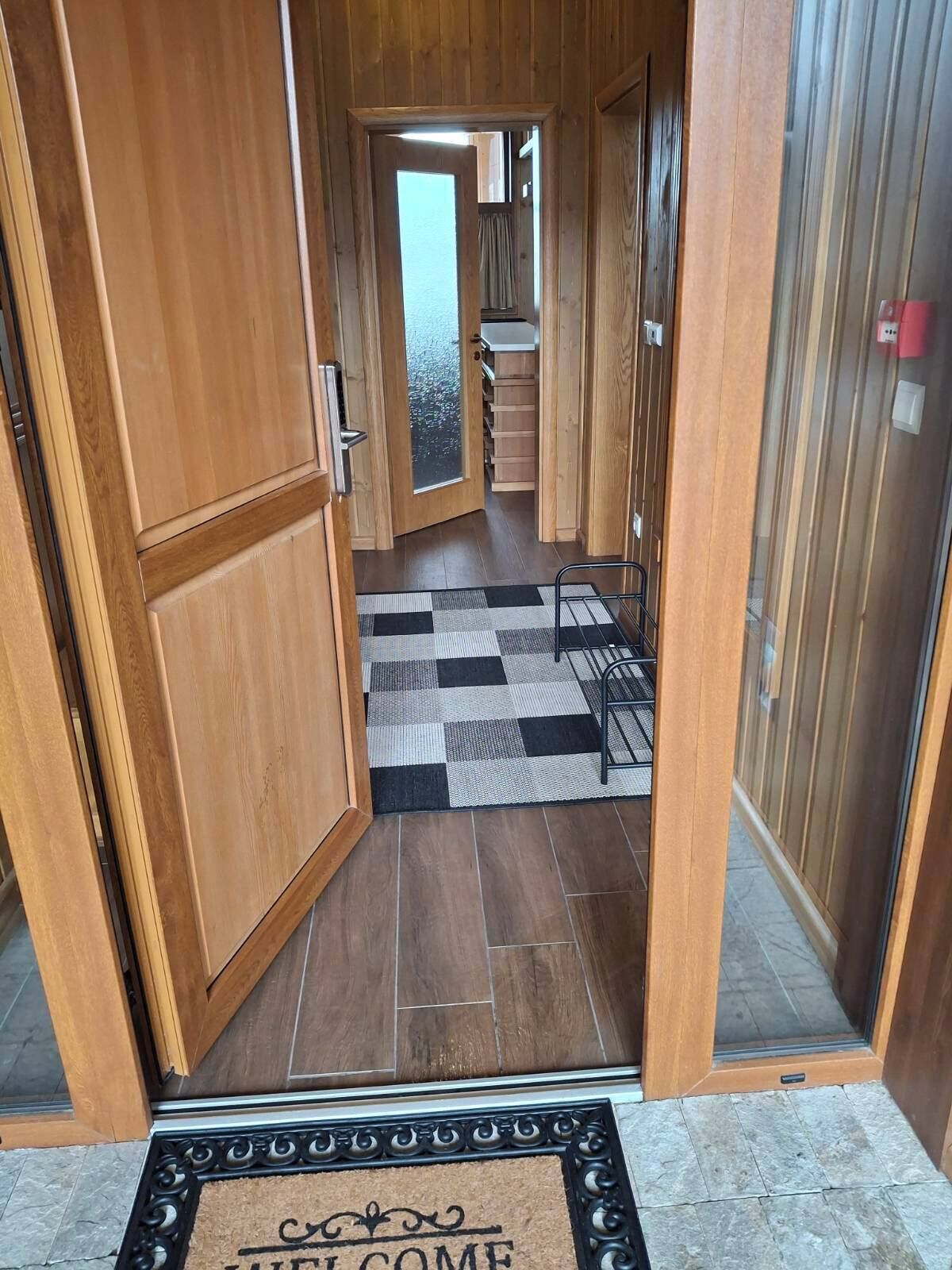 Pasillo interior de madera con felpudo a cuadros, un estante metálico y vista a una zona de cocina.