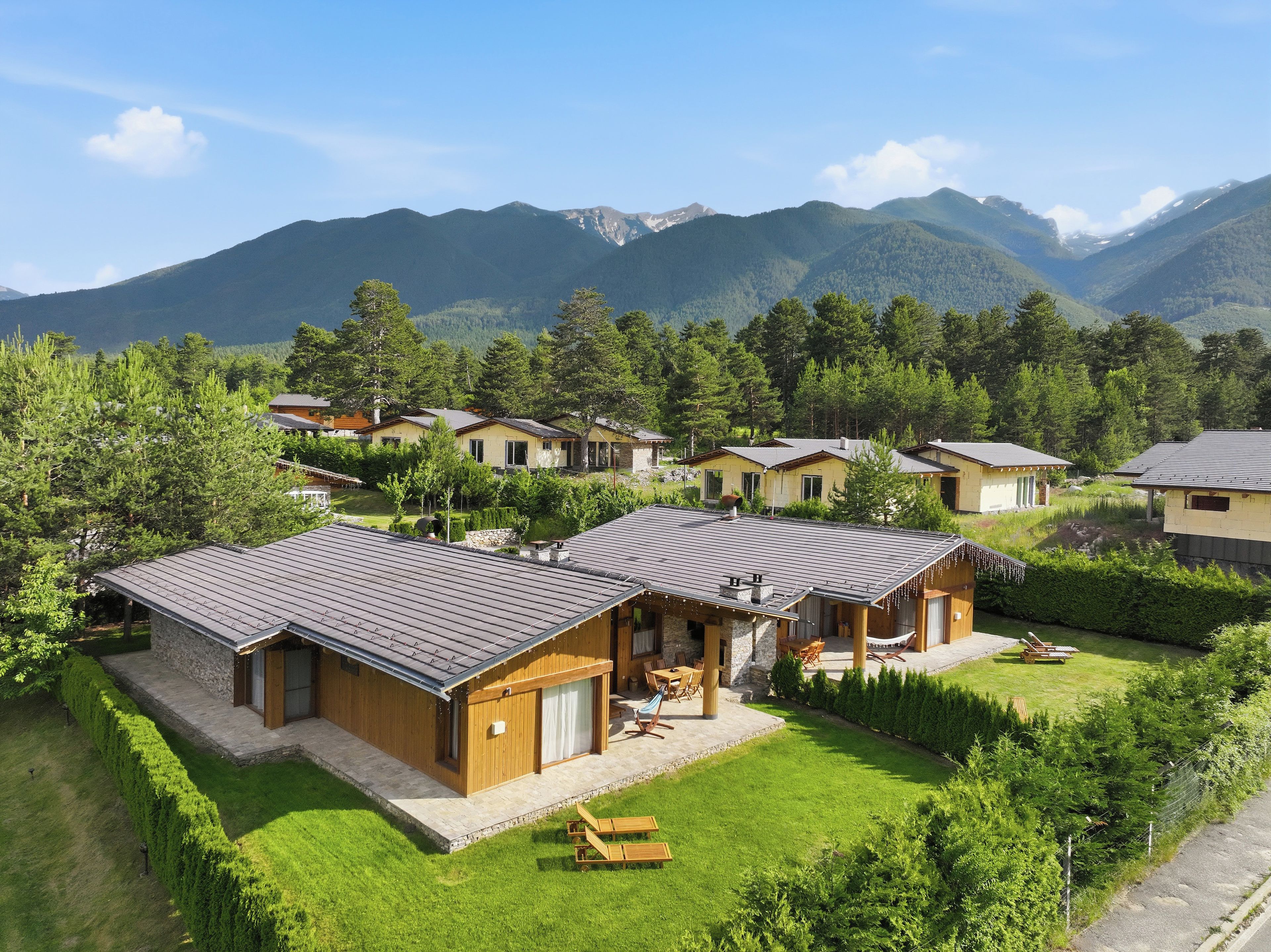 A cluster of wooden cabins with sloped roofs, surrounded by lush greenery and mountains in the background.