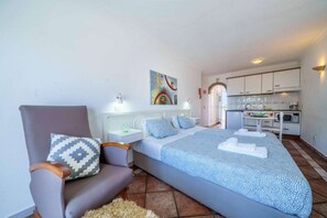Un dormitorio con cama, una silla, una mesita de noche, un cuadro y una kitchenette al fondo.