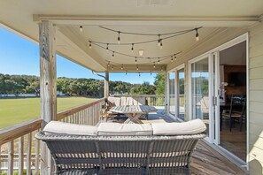 A covered outdoor patio with wicker furniture, string lights, and a view of a grassy field. - The Sand Wedge: Dogs Allowed | Private Saltwater Pool (Kitty Hawk)