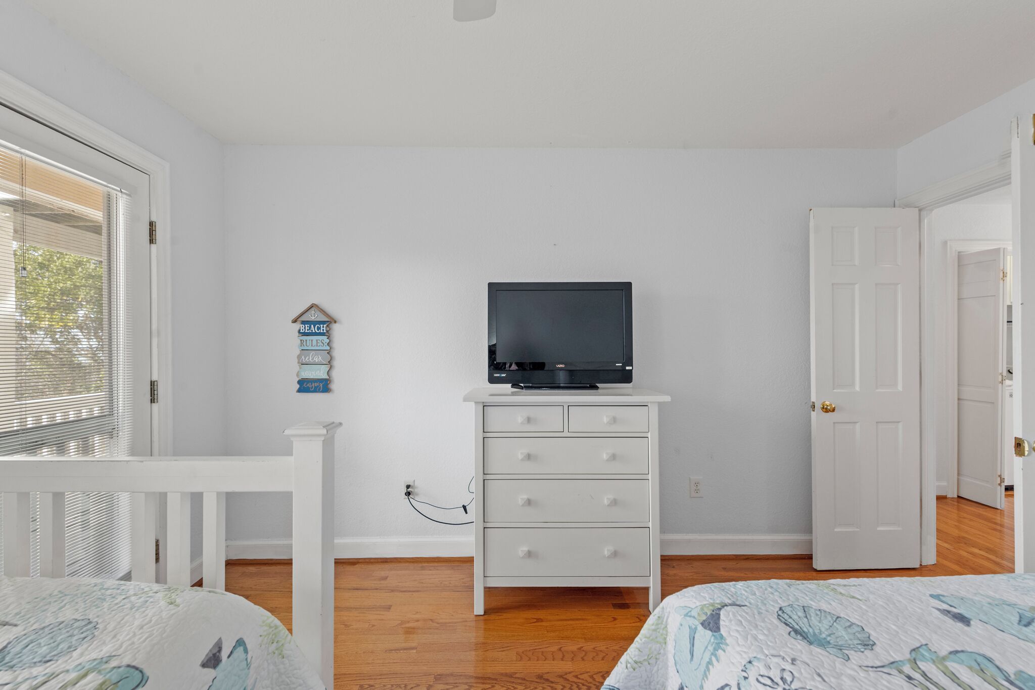 Une chambre avec une commode blanche, un téléviseur posé dessus, un lit recouvert d’une couvre-lit à motifs floraux et une porte donnant sur une autre pièce.
