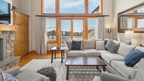 Ein Wohnzimmer mit Kamin aus Stein, einem großen Fenster mit Vorhängen, einer Couch und einem Fernseher.