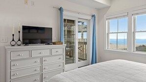 Una camera da letto con un letto, una TV su un comò, un balcone vista spiaggia e una finestra con tende.