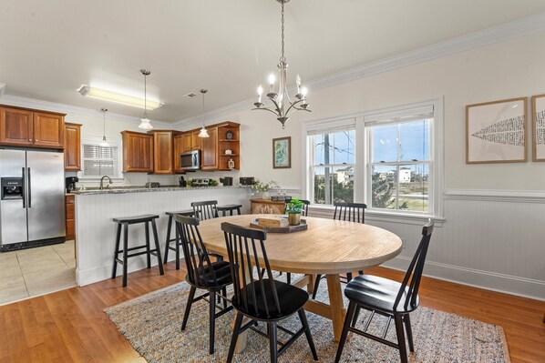 A modern kitchen with wooden cabinets, a stainless steel refrigerator, a round wooden dining table with six chairs, and a chandelier. - Libby's Keep: Oceanfront | Elevator (Rodanthe)