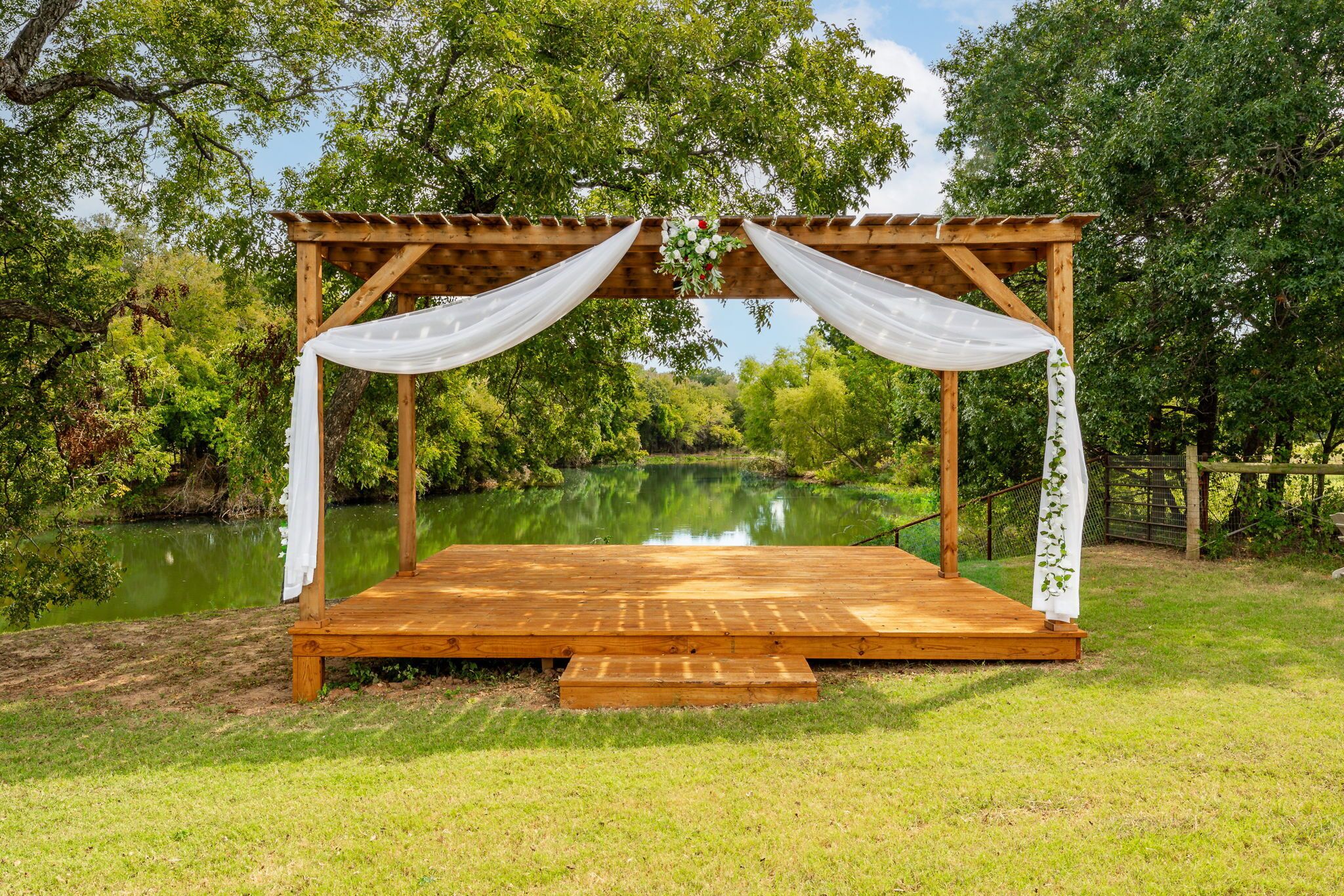 A wooden gazebo with white drapes, a wooden platform, and a view of a lake surrounded by trees.