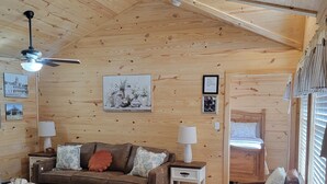 Ein gemütliches Blockhaus-Interieur mit Holzbalkendecke, einem braunen Ledersofa, einem Esstisch mit Tellern und einem Bett mit Fenster.