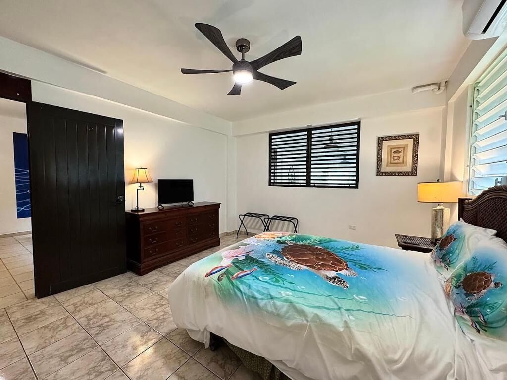 Un dormitorio con cama, ventilador de techo, cómoda, televisor y ventana con persianas.