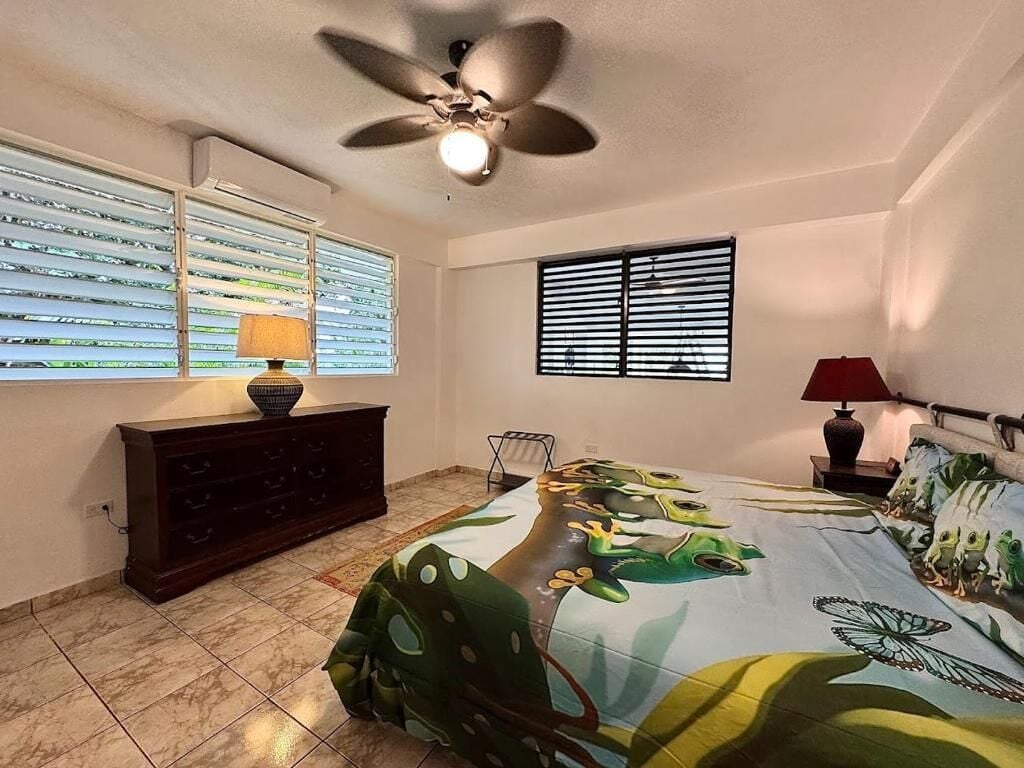 A bedroom with a bed, a ceiling fan, a window with blinds, a dresser, and a lamp.