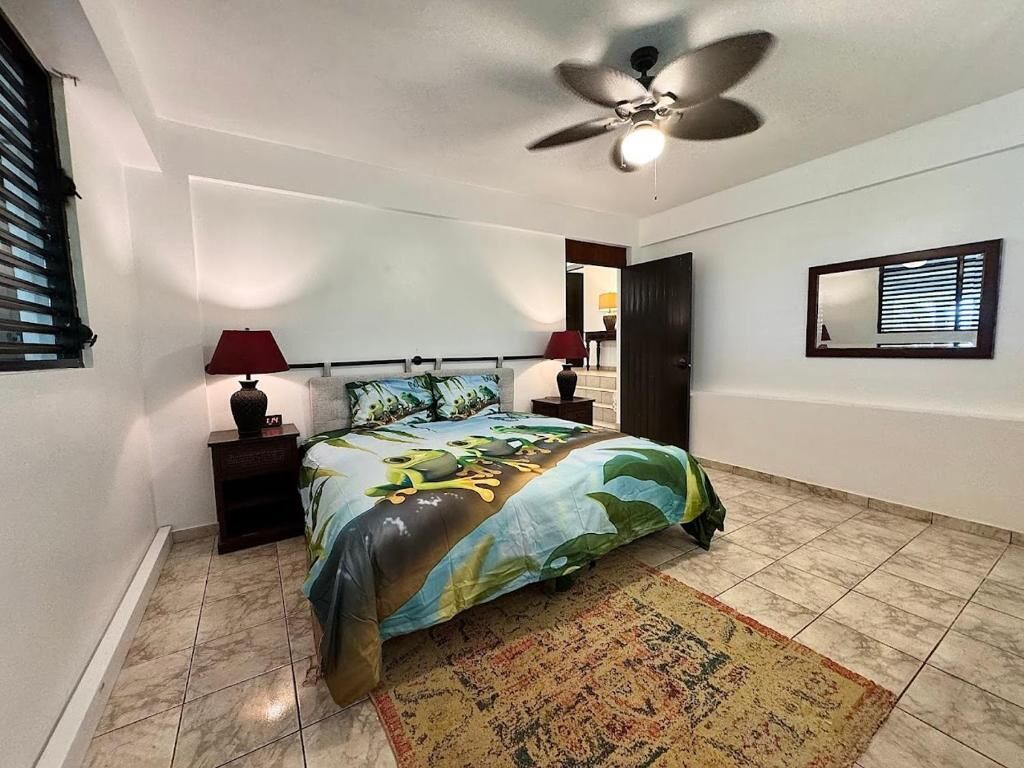 Un dormitorio con cama, dos mesitas de noche, ventilador de techo, ventana con persianas y un espejo en la pared.