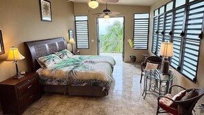 A bedroom with a bed, two ceiling fans, a nightstand, a chair, and a window with shutters. - Vieques BioBay Bed and Breakfast (Vieques)