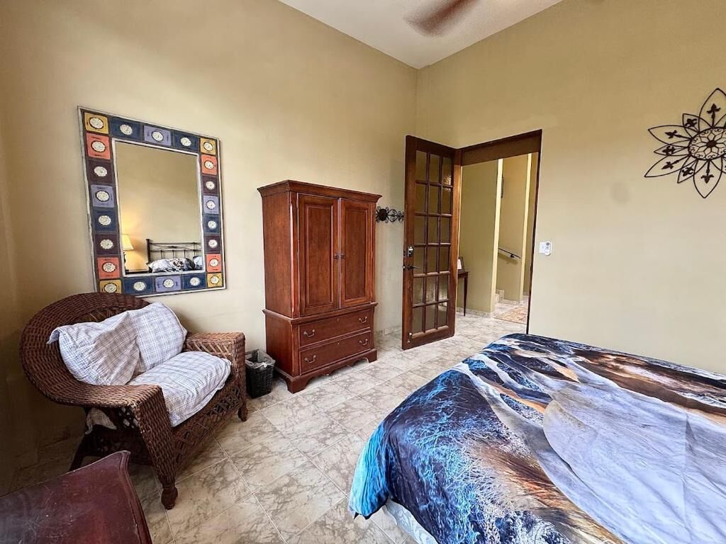 Un dormitorio con una cama, una silla de mimbre, un armario de madera, un espejo y una puerta que da a otra habitación.