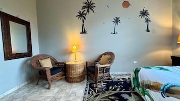 A room with a bed, two wicker chairs, a small table with a lamp, a patterned rug, and wall art of palm trees and birds.
