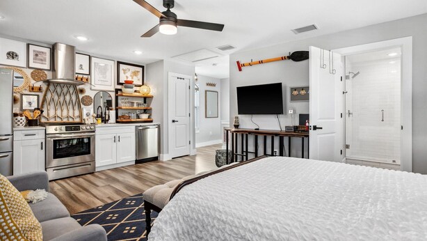 Un dormitorio moderno con una cama grande, ventilador de techo, televisor y una kitchenette al fondo.