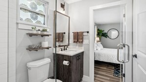 Un baño moderno con inodoro, lavamanos y ducha. Se ve una cama a través de una puerta abierta.