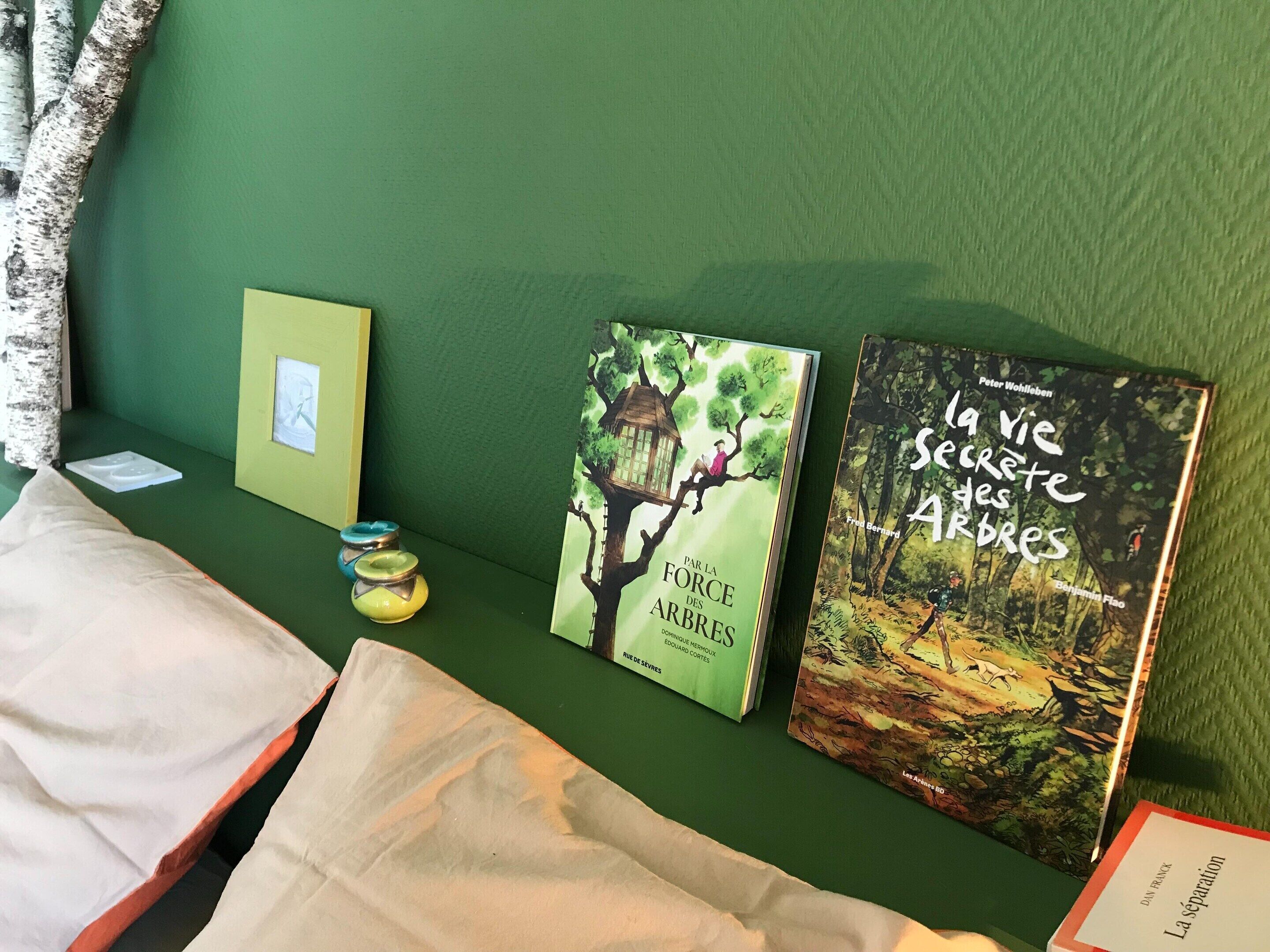 Una habitación con pared verde, cama con ropa de cama blanca y naranja, y dos libros titulados 'PAR LA FORCE DES ARBRES' y 'La vie secrète des ARDRES'.