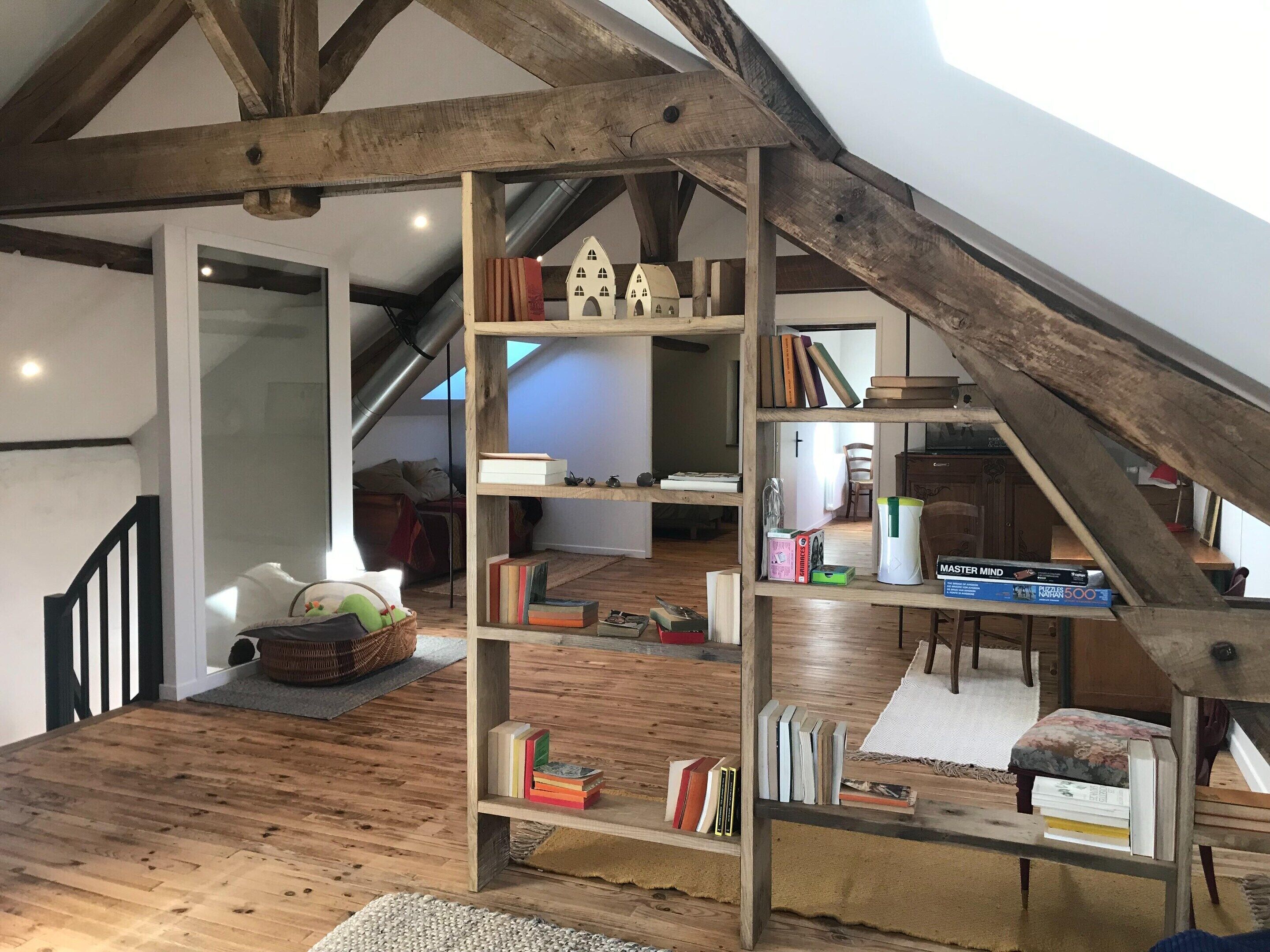 Une chambre à coucher de style « loft », avec des poutres apparentes, un plafond en pente et une bibliothèque intégrée remplie de livres et d’objets décoratifs.