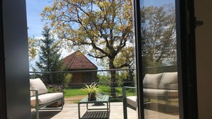 Vista da una stanza con porta a vetri, su un'area esterna in legno con mobili da giardino, un tavolo con una pianta e un edificio con tetto rosso sullo sfondo.