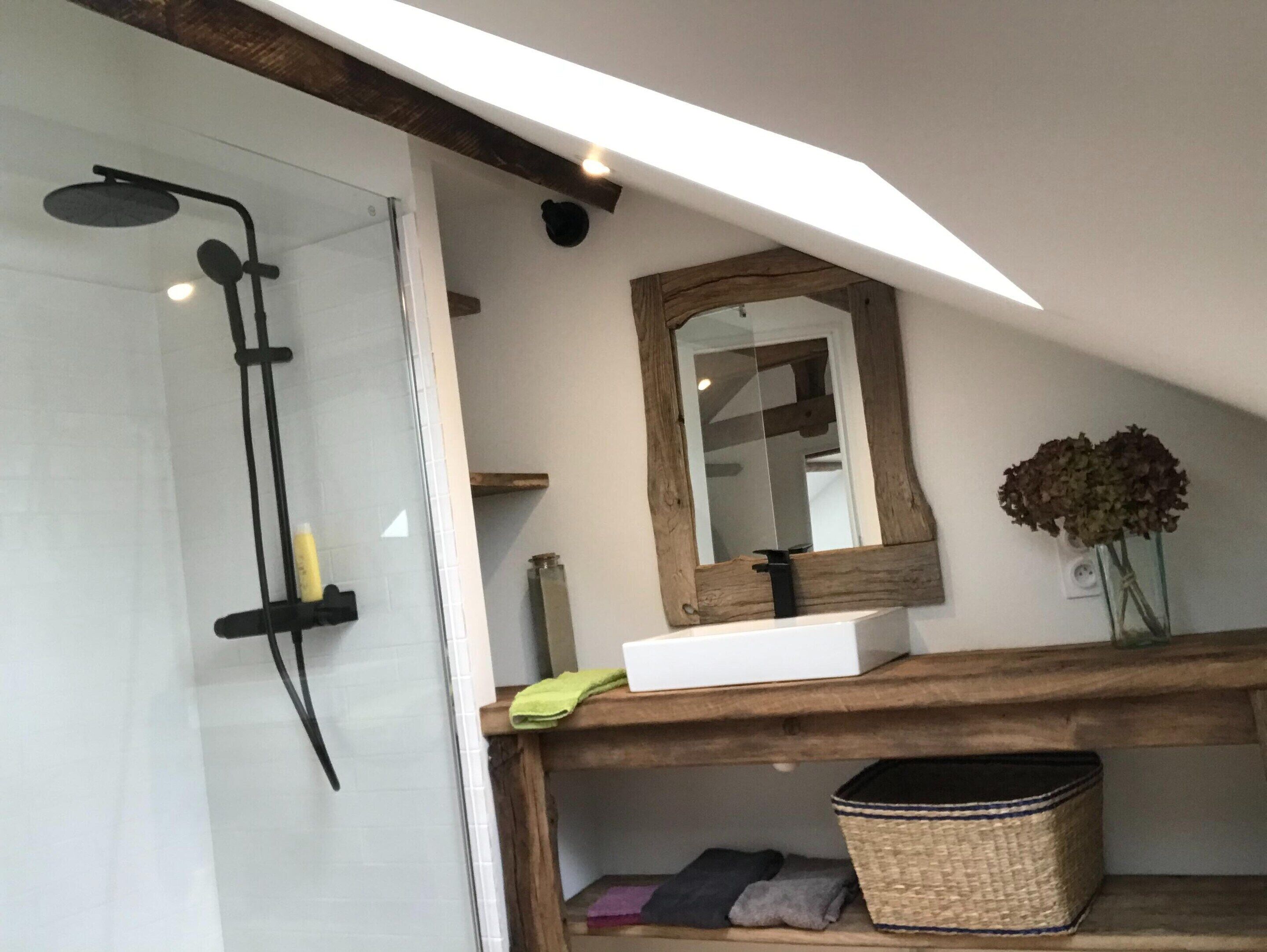 A bathroom with a glass shower enclosure, a wooden shelf with a mirror, and a vase with flowers.