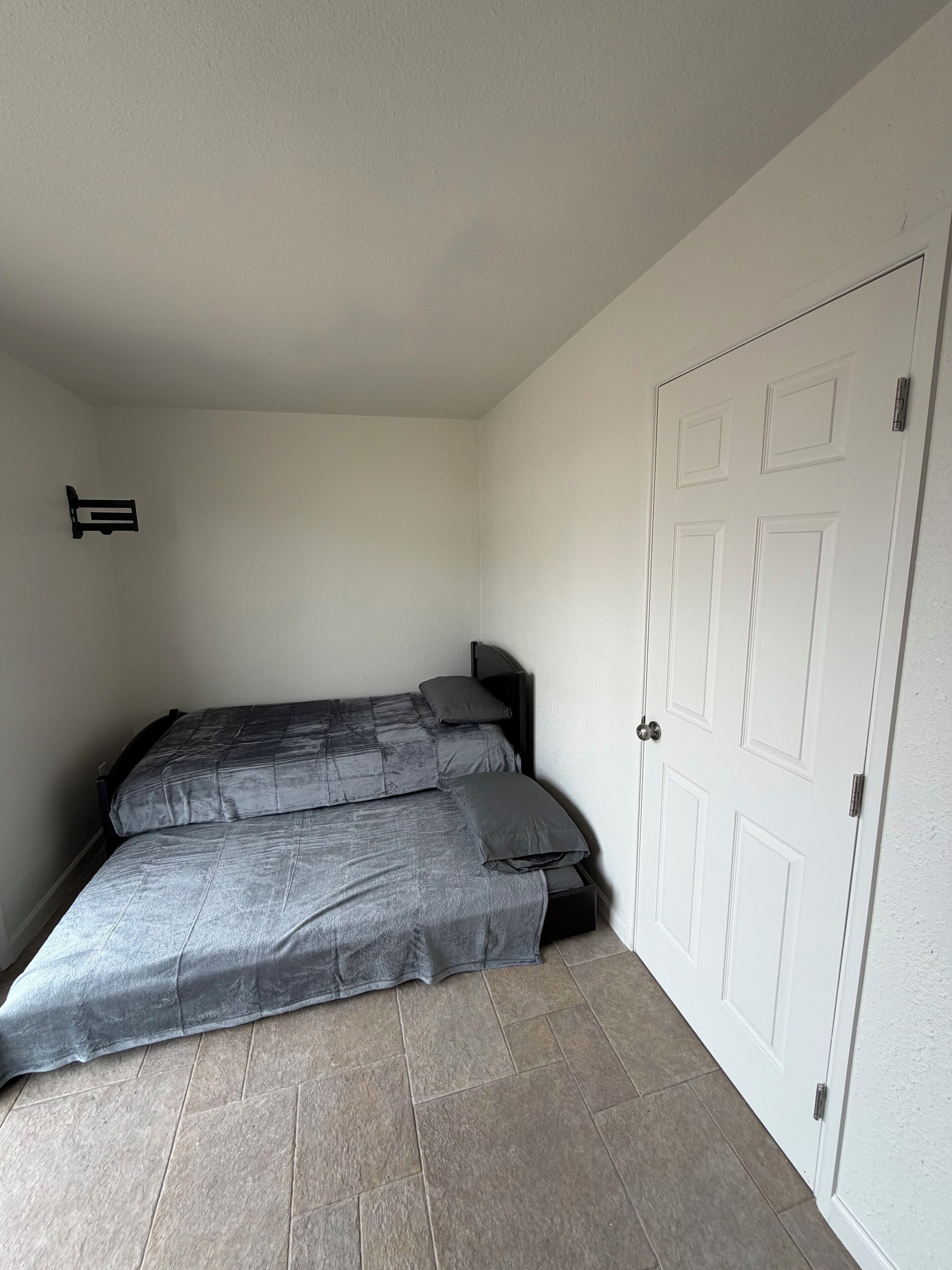 A small, single-bed room with a plain white door and a small shelf on the wall.