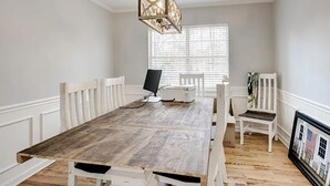 Een eetkamer met een houten tafel, witte stoelen, een hanglamp en een raam met luxaflex.