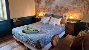 Een kamer met een groot bed, twee nachtkastjes met lampen, een houten stoel, en een muurschildering van palmbomen en vogels.