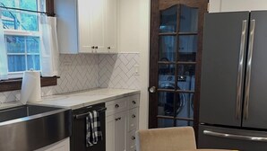 Eine moderne Küche mit weißen Schränken, einer Edelstahlspüle und einem schwarzen Kühlschrank. Ein Esstisch mit Stühlen und eine Schüssel mit Obst stehen darauf.