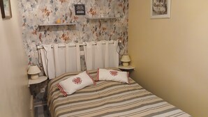 A bedroom with a striped bedspread, two pillows with heart designs, a radiator, and a small shelf with decorative items. - Small house located in a village 15min from Langeais (Hommes)