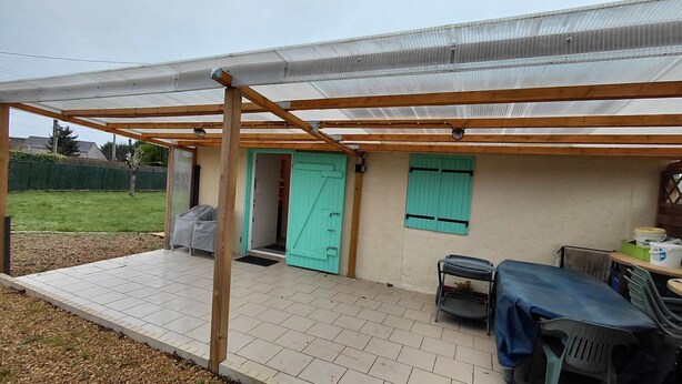 A covered patio area with a tiled floor, a wooden pergola, a teal door, and a table with chairs. - Small house located in a village 15min from Langeais (Hommes)