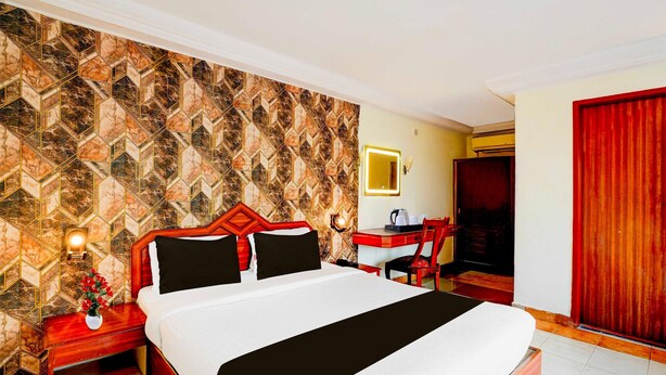 A hotel room with a large bed, a desk with a chair, a mirror, and a red door. - Deluxe Olive Star (Mysore)