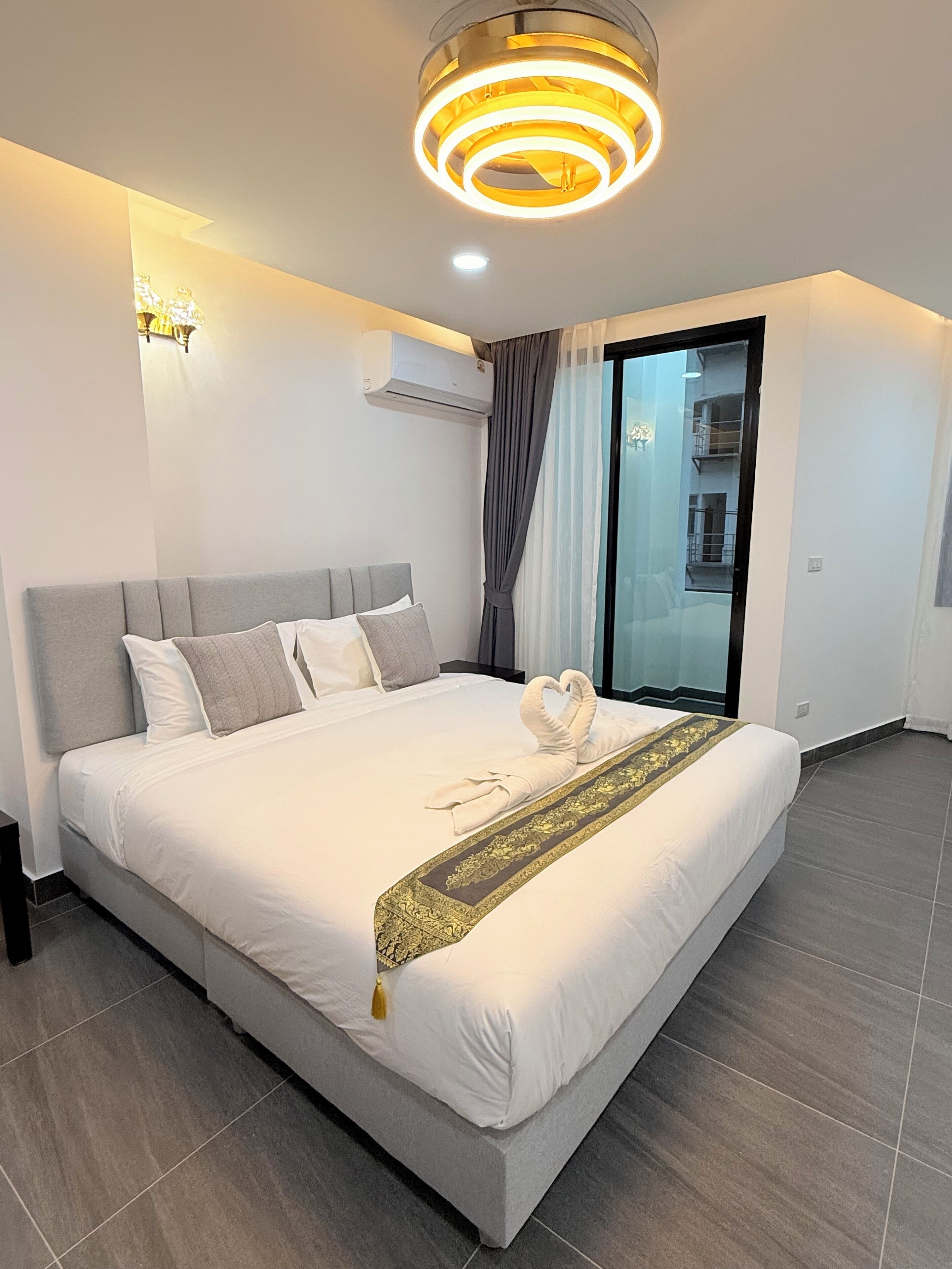 A modern hotel room with a large bed, a decorative towel arrangement, a ceiling light fixture, and a view through a glass door.