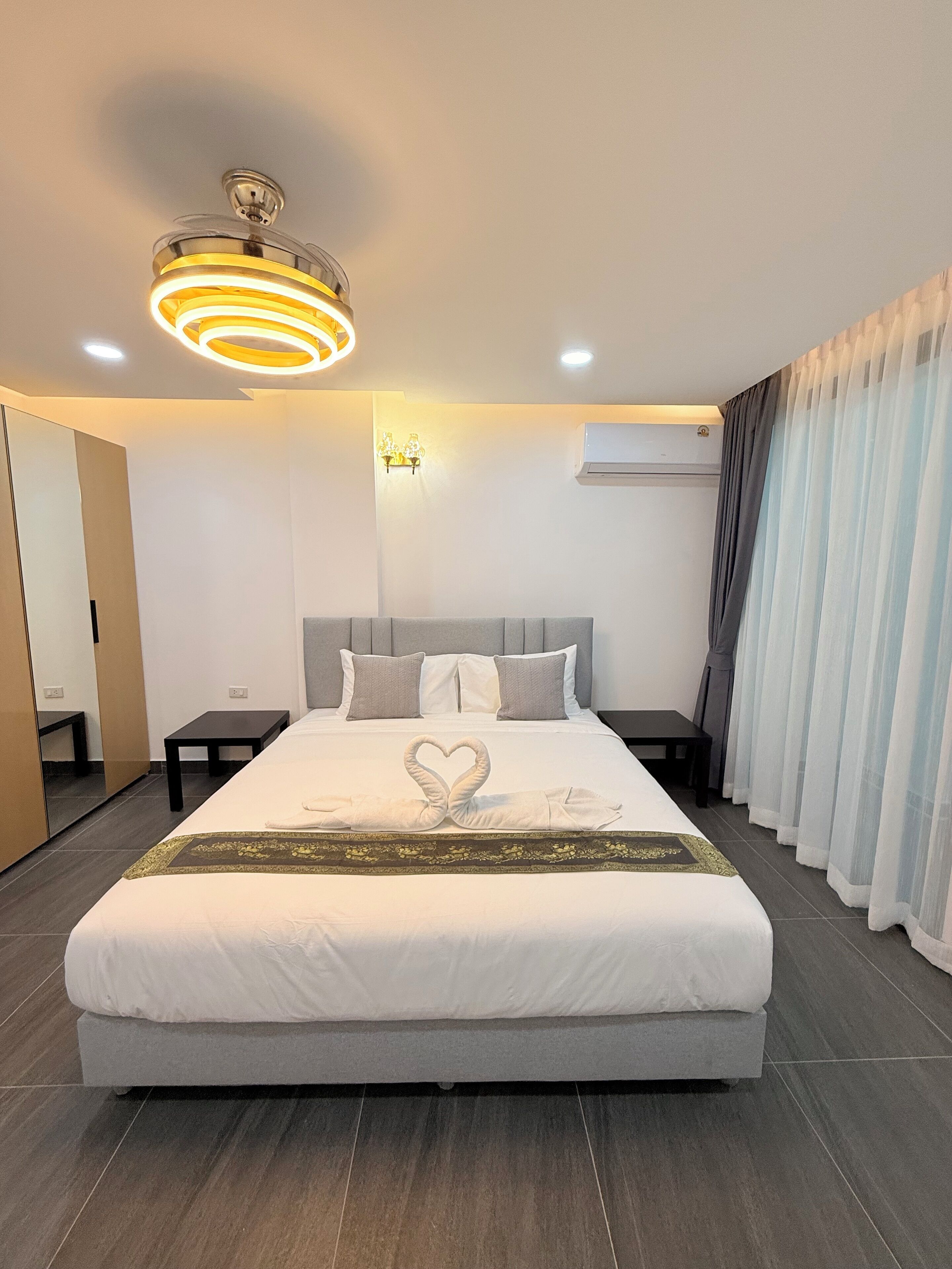 Une chambre d’hôtel moderne dotée d’un grand lit, d’un luminaire au plafond, d’un climatiseur fixé au mur et d’une fenêtre avec des rideaux légers.