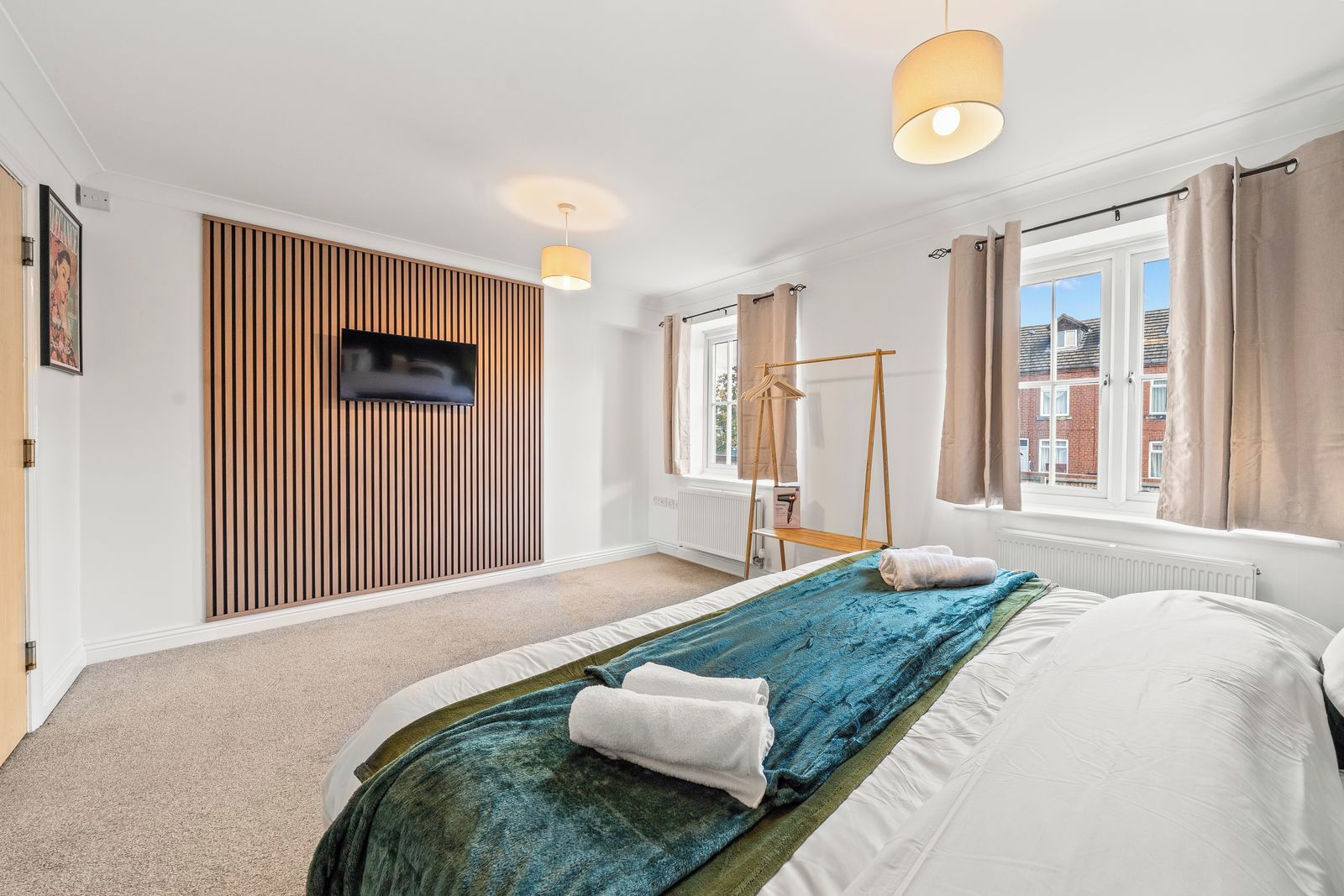 Una camera da letto con un letto, una televisione montata su un pannello di parete in legno, una finestra con tende e un termosifone.