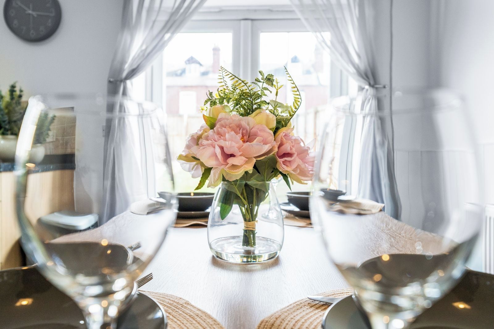 Une table à manger ornée d’un vase de fleurs, de verres transparents et d’une horloge au mur.